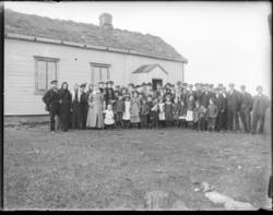 Gruppebilde tatt utenfor en lav bygning med torvtak. Menn, k