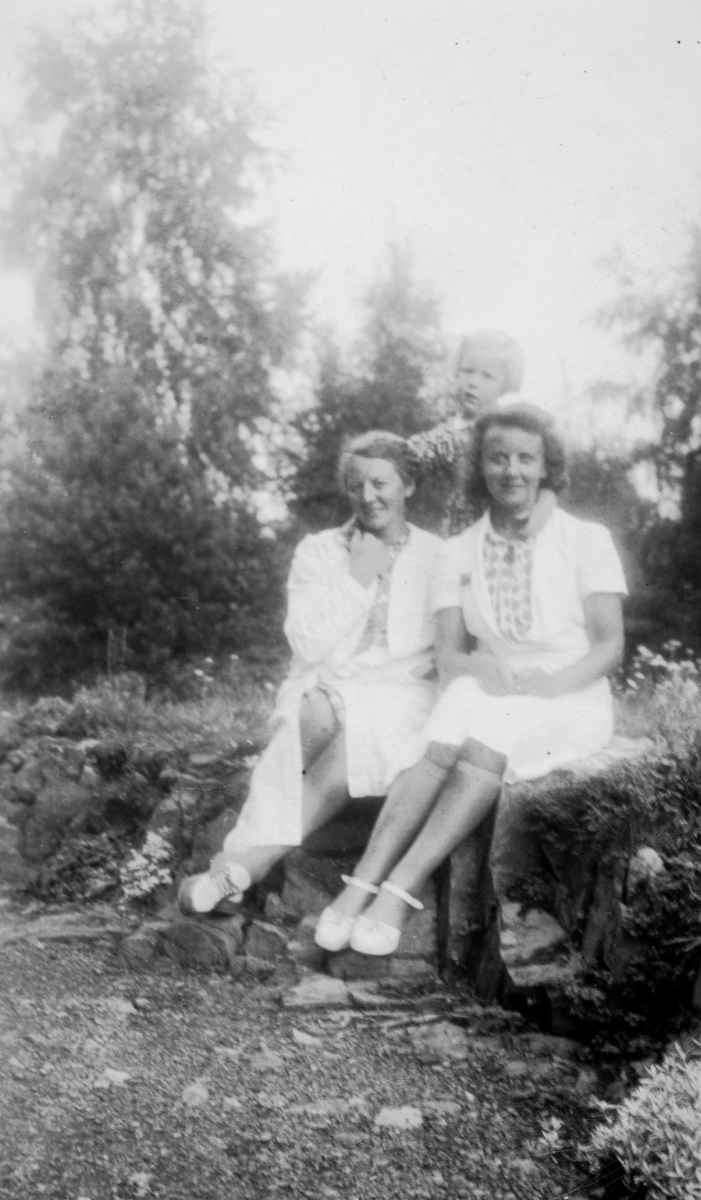 To damer sitter på bakken Bak står en liten pike. Det er trær rundt dem.Damene har lyse kjoler. Olga og Gull.