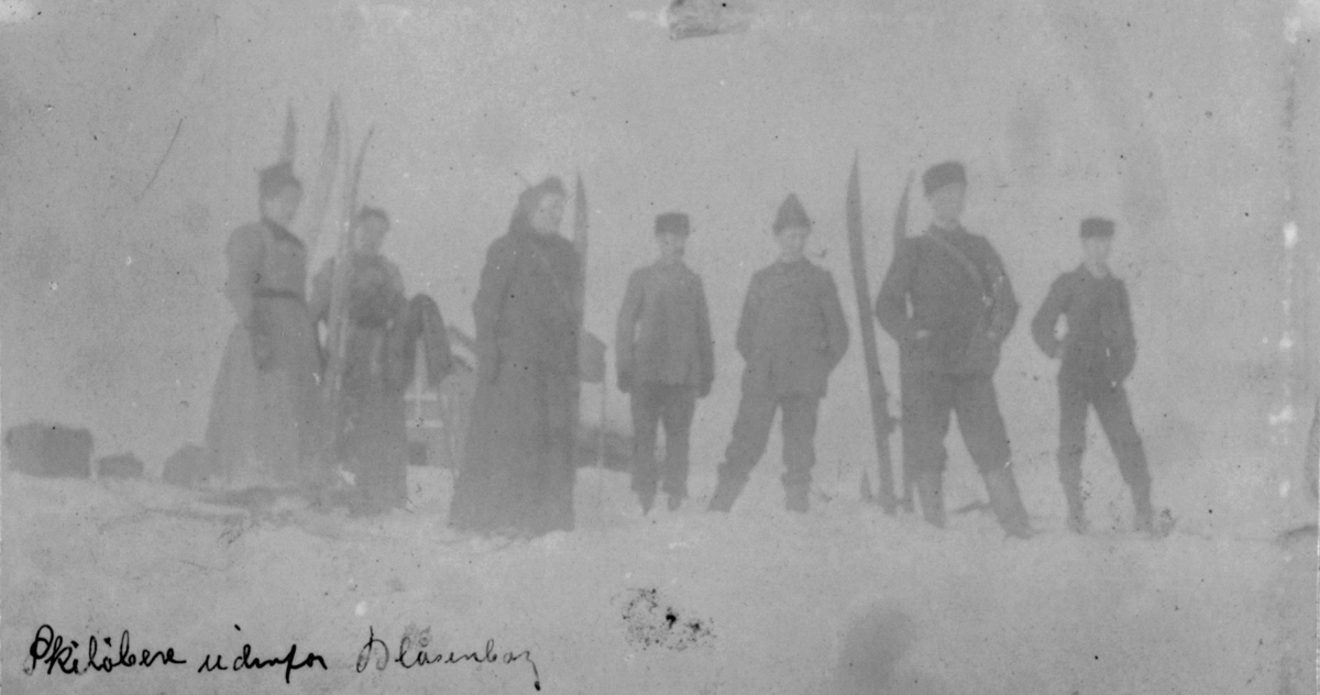 Skiløpere utenfor Blåsenberg. Fire menn og tre damer står i snøen. Damene har side kjoler. Ski står satt ned i snøen.