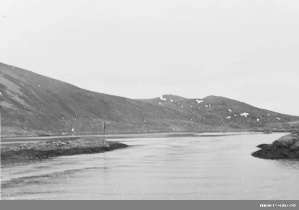 Naturbilde. Tatt utover fjorden i Tufjord. En åskam strekker seg utover fra venstre billedkant. Sjøen ligger rolig. Det er overskyet.Et skjær til venstre i bildet.