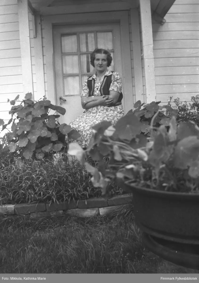 Astrid Lindseth sitter ved inngangsdøra til gården Mikkelsnes. I forgrunnen ser vi frodige hageblomster, bl.a. er blomkarsen lett å kjenne igjen