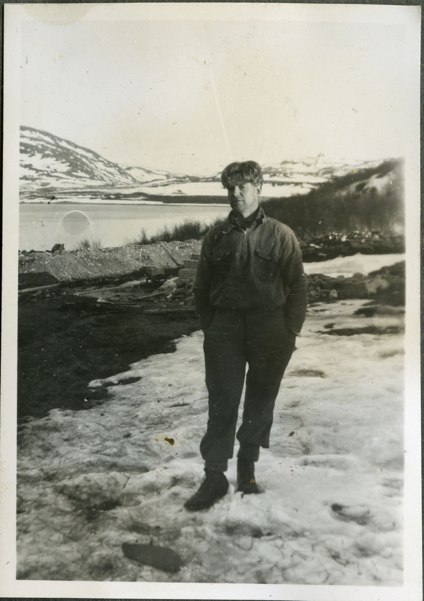 Fotografi av en ukjent mann på skiferbruddet i Friarfjord. Mannen er trolig en arbeider på stedet. Han er kledd i skjorte, bukse, sko og et skjerf rundt halsen. Bak ham kan man se steiner og byggeredskaper. I bakgrunnen ser man havet og fjell. På bakken ligger det snø.