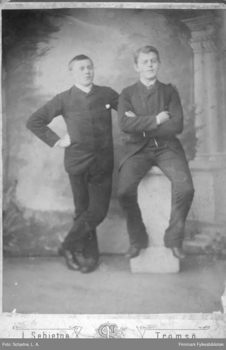 Portrett av to unge menn i mørke dresser og sko. Han til venstre på bildet står med beina i kryss. Mannen til høyre på bildet sitter på en liten søyle med armene i kryss. Veggen bak dem har tapet med et bildemønster.