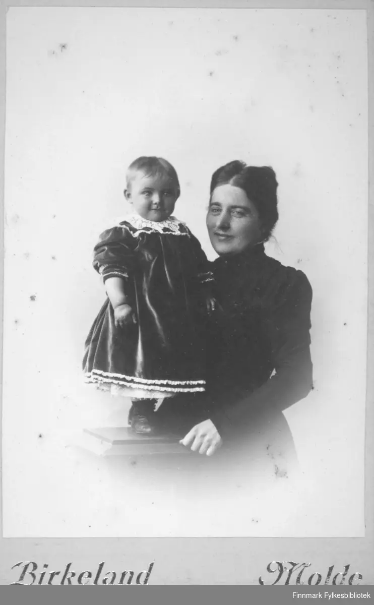 Portrett av en kvinne og et barn. Kvinnen har en mørk overdel på seg. Hun holder armen rundt barnet som står på en søyle og er iført mørk kjole med hvit krage.