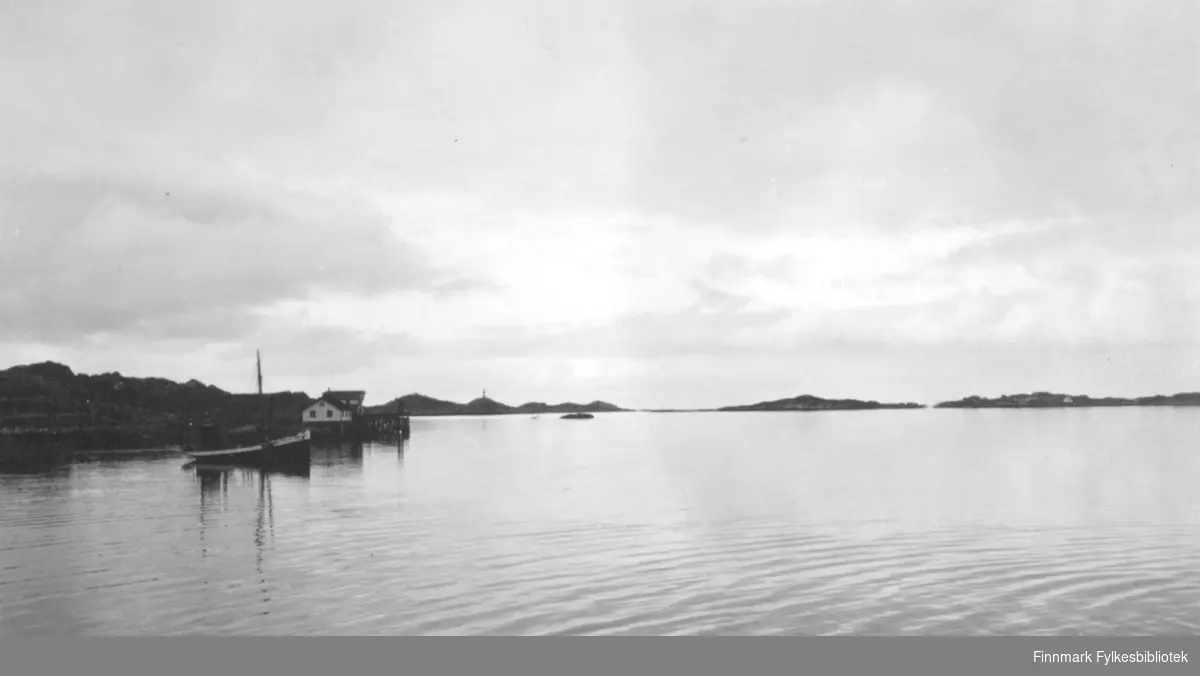 En sommerdag på Ingøy. Blikkstille hav og skyet. En båt ligger fortøyd på havna og et hvitt hus ligger i sjøkanten. Et sjømerke står på en liten topp og en holme ses litt til høyre for sjømerket. Flere små øyer ses mot horisonten til høyre på bildet.