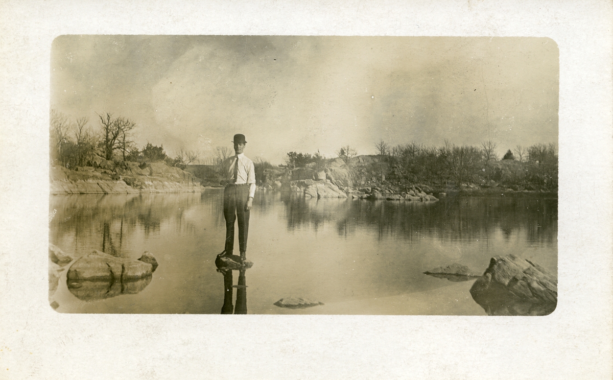 Mann avbildet på stein i vannkanten. Mannen er iført skjorte og slips  og med hatt. Dette er et prospektkort.