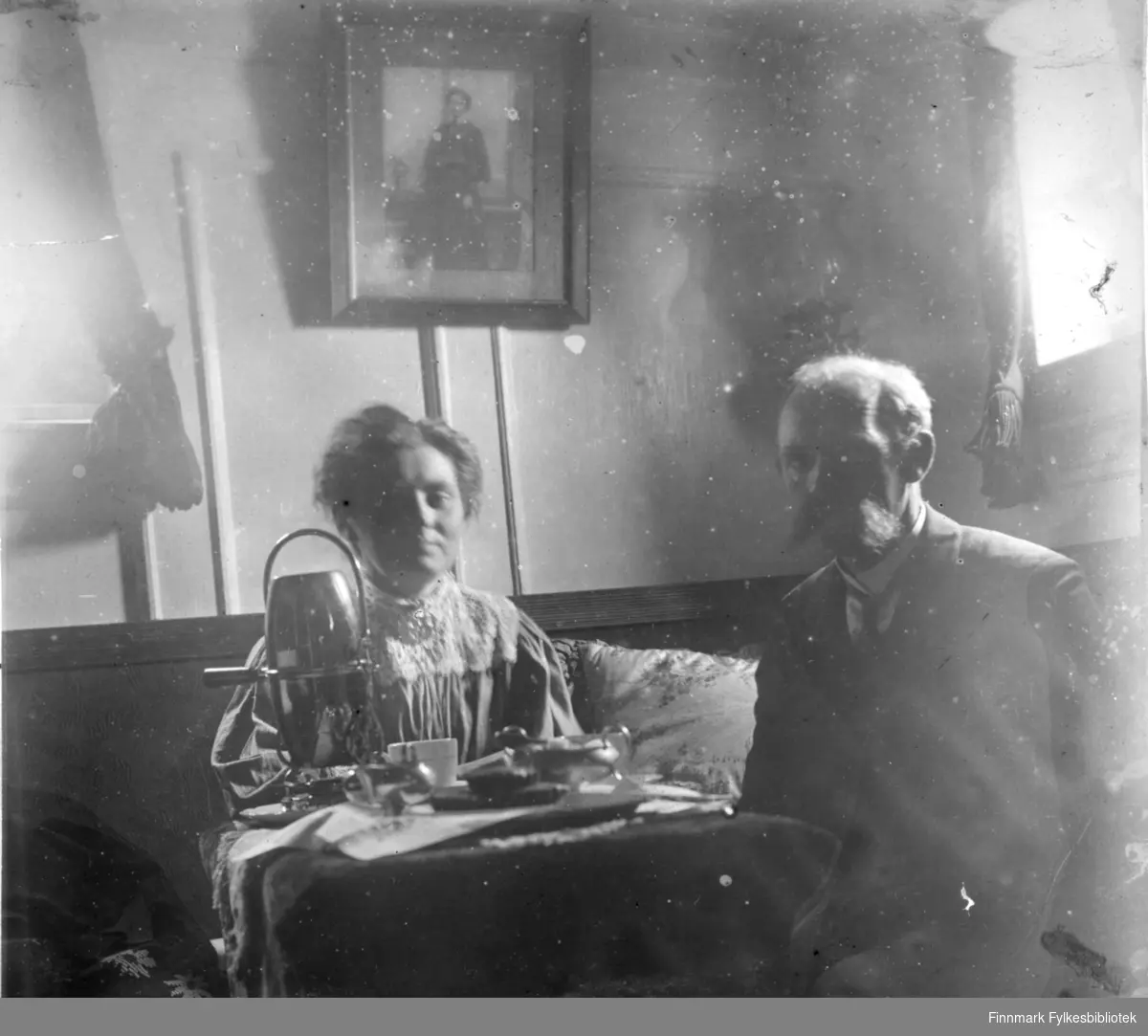 En dame og en mann sitter ved et lite, rundt bord. Det er Theodor Augensen, som tidligere var havnefogd i Hammerfest. Ved siden av han sitter hans kone. Det ligger en heklet duk på bordet med kopper, tefat og en kaffekjele på stativ. Damen har en ganske mørk overdel med et sjal rundt halsen. Mannen har dressjakke med hvit skjorte og slips. På veggen bak henger et fotografi og i sofaen står en sofapute ved siden av damen. To vinduer vises på begge veggene og det har ganske mørke gardiner.