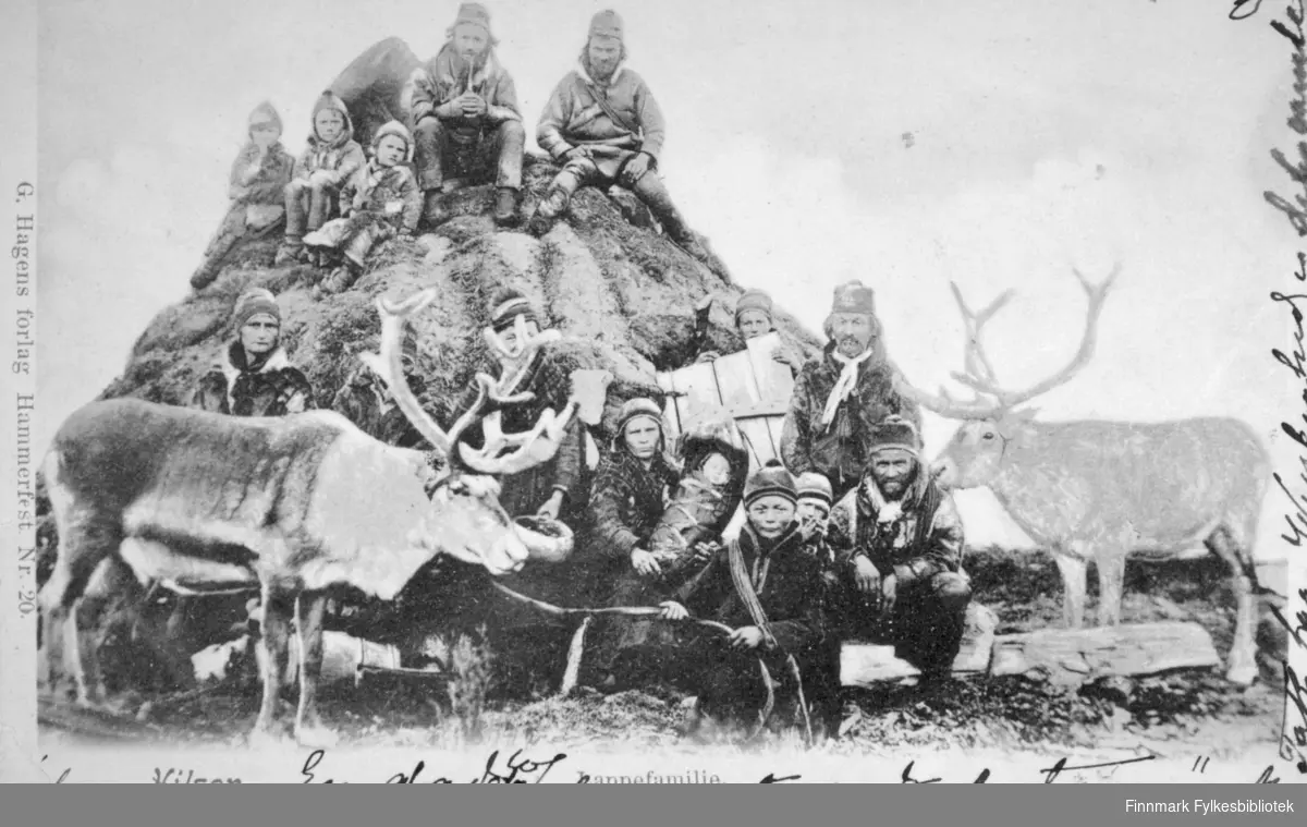 En samisk familie poserer ved og på en gamme. Denne familien flyttet til Kvaløya utenfor Tromsø på sommeren og reiste til Karesuando i Sverige på vinteren. Bildet er tatt i 1880-90-årene. To menn og tre barn sitter på toppen av gammen. Ni personer sitter foran gammen. Flere barn, deriblant et spedbarn, sitter midt på bildet. To reinsdyr med horn står på hver side av bildet. Alle sammen har hodeplagg på og de fleste har også pesk. Spedbarnet er innpakket og en ung dame holder barnet i fanget. Den unge gutten helt foran på bildet har en lasso hengt over skuldrene. Det er gress/lyng på bakken foran på bildet og det er mye skyer på himmelen.
