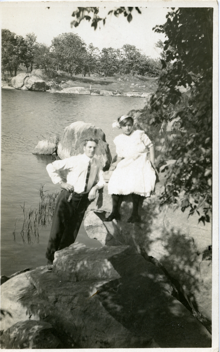 Kvinne og mann avbildet ved vann. Kvinne er iført kjole og mannen med skjorte og slips. Postkort.