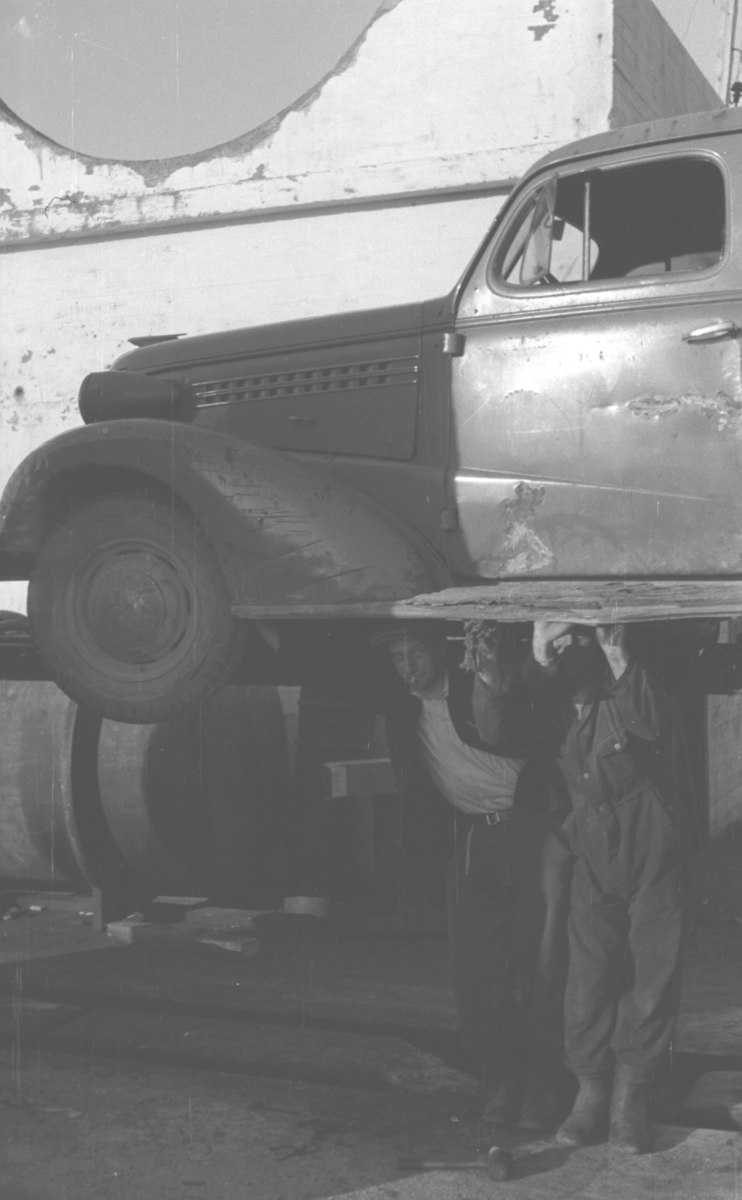 En Chevrolet, årsmodell 1938,  er heist opp på en bukk og to menn står under den. Personer og sted er ukjent.