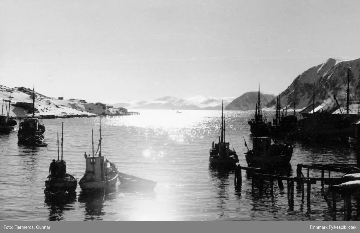 Sola speiler seg i sjøen en sen viterdag. Flere båter ligger fortøyd i havna og en del snø ligger i terrenget. Stedet er ukjent, men det kan være fra Honningsvåg.