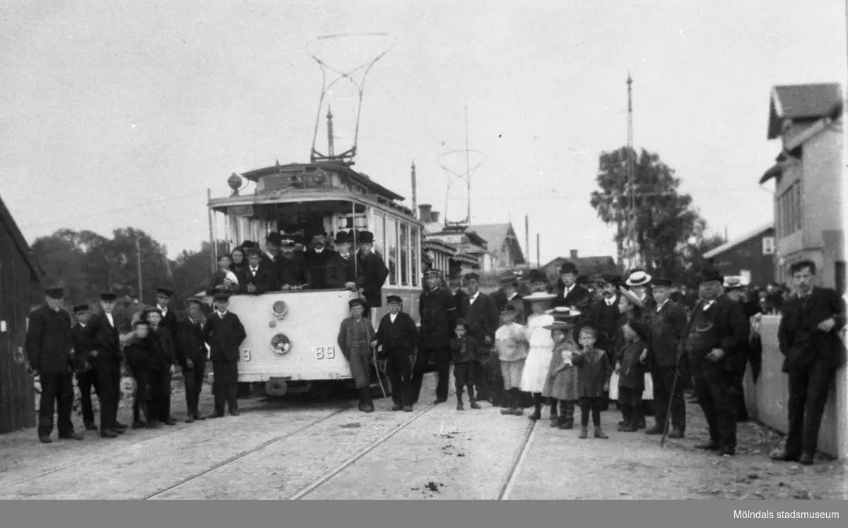 Spårvagn med människor runtomkring. Det var en stor dag den 14 augusti 1907, då den elektriska spårvägslinjen Göteborg - Mölndal togs i bruk. Den dagen var arbetarna fria, och de hade mangrant samlats utefter vägen, när den första spårvagnen avgick från Mölndalsbro. Flaggorna i hela samhället och inne i staden var i topp, och det blev ett väldigt hurrarop och viftande, då den första vagnen äntligen syntes på avstånd, i vilken representanter för Mölndals kvarnby samt spårvägsstyrelsen åkte.