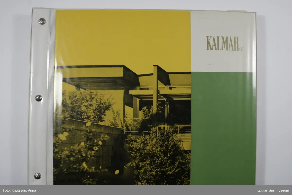 KLM 33297. Tapetkatalog. Plastpärm i vitt, gult och grönt med ett fotografi av en byggnad på framsidan. Tapetkatalog för Kalmar Nya Tapetfabrik, innehåller många olika prover av tapeter från 1970-talet. Datering: 1970-talet.