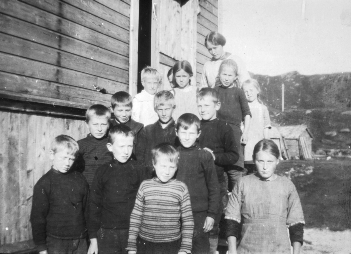 "Småskolen. Breivik 1916". Barn står på trappen til en skole i Breivik. Jentene er kledd i kjoler. Guttene har på seg gensere. I bakgrunnen kan man se et lite trehus og fjell.