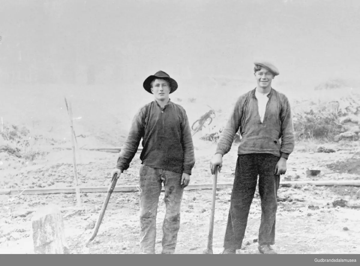 To unge menn utendørs, står med spade og hakke, i arbeid med bygging av jernbane?, det er høst, utydelig bakgrunn.