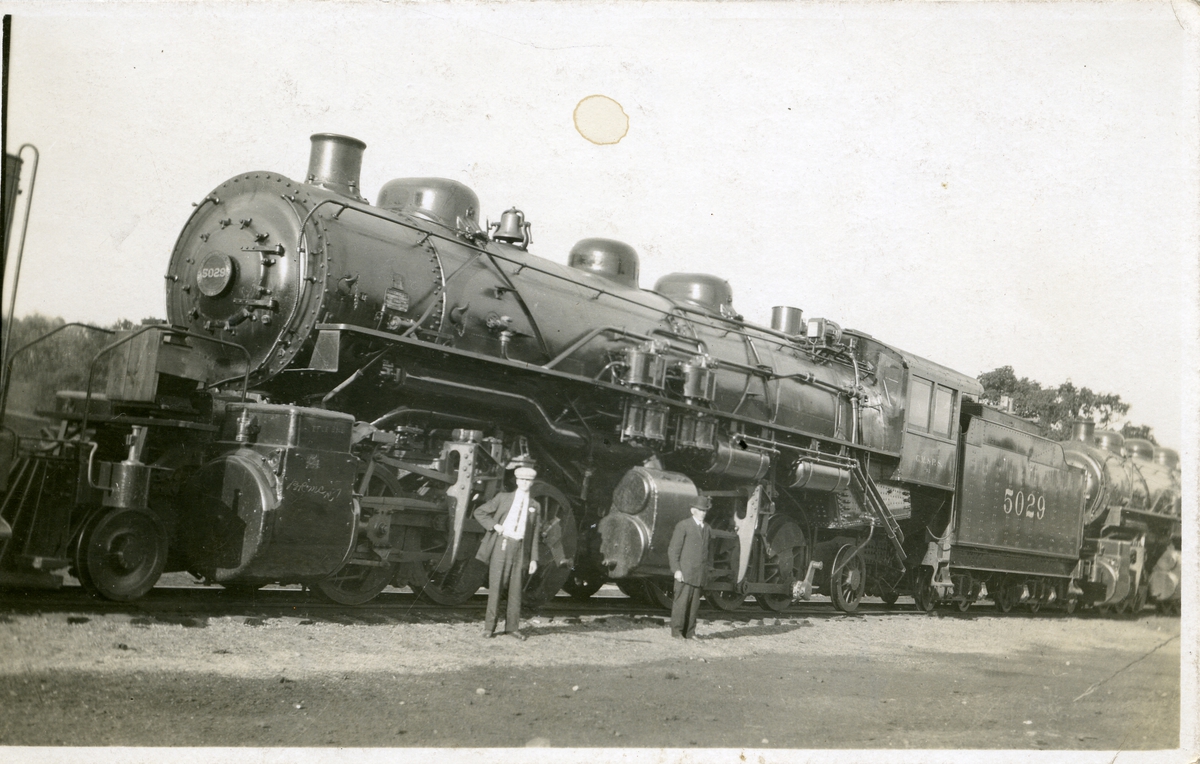 Bildet viser to personer foran et lokomotiv.
Dette er et postkort.