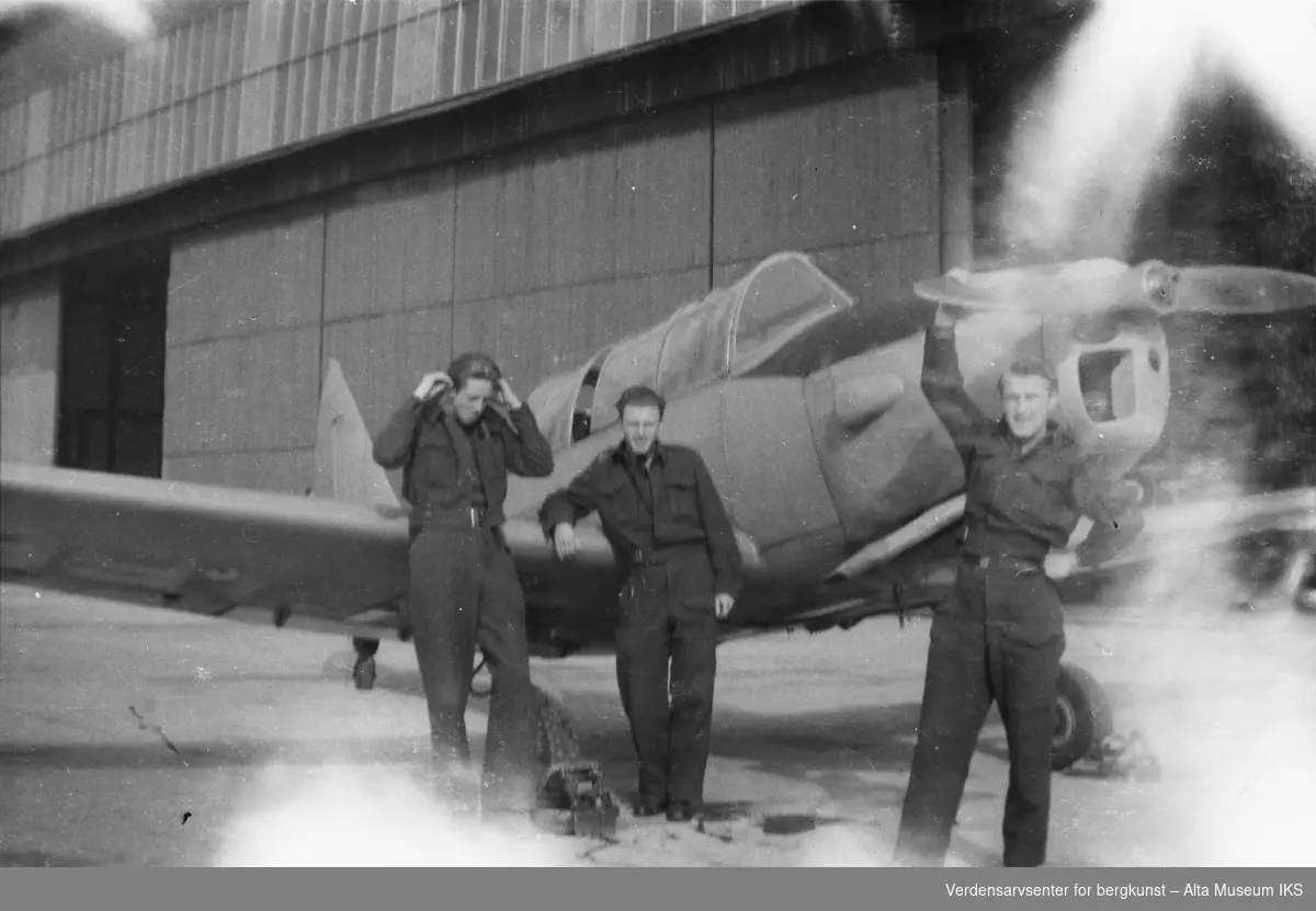 Tre unge menn i uniform står ved et militærfly ved en hangar.