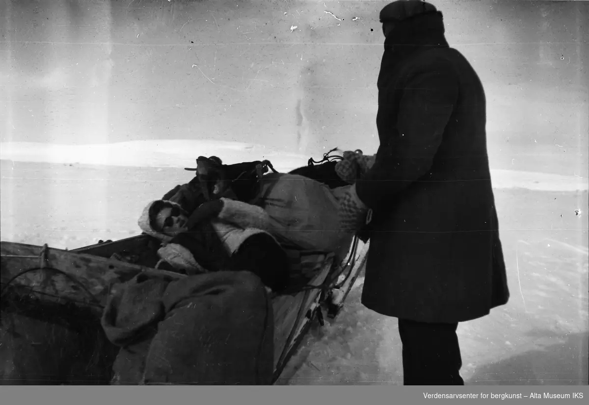 Erna Jøraholmen slapper av i sleden, og en mann står ved siden av med ryggen til kameraet. Bildet er fra en fjelltur med hest og slede.