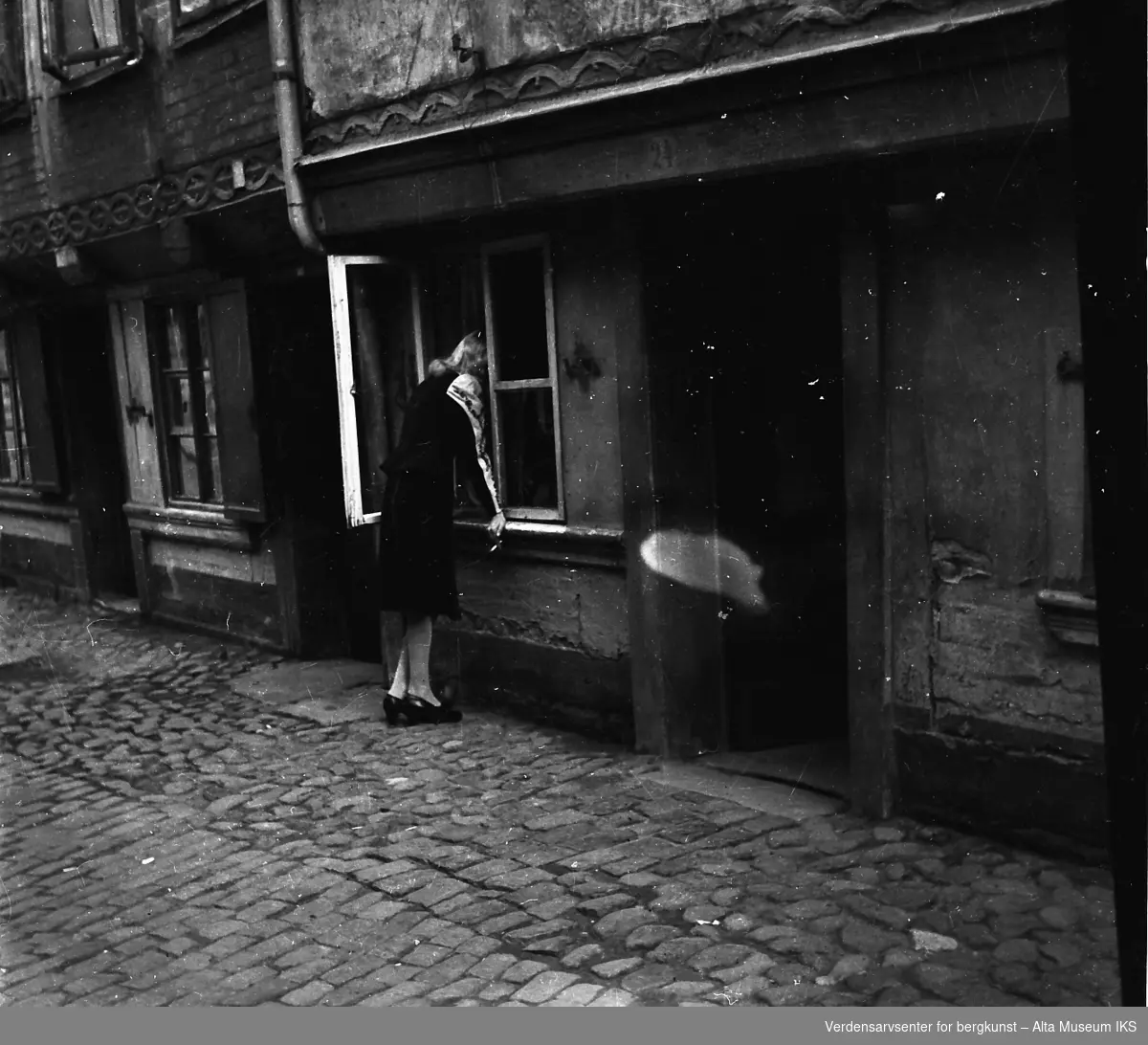 En kvinne stikker hodet inn et vindu. Hun står i en gate med brostein.