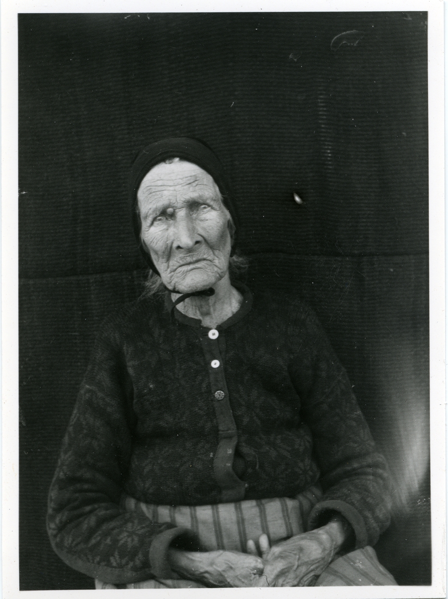 Portrett i halvfigur av eldre dame. Hun er kledd i strikkejakke og forkle og har en lue/kyse på hodet.