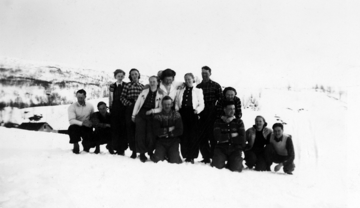 Gruppebilde av menn og kvinner, tatt ute i snøen, med fjell og en bygning så vidt synlig i bakgrunnen.