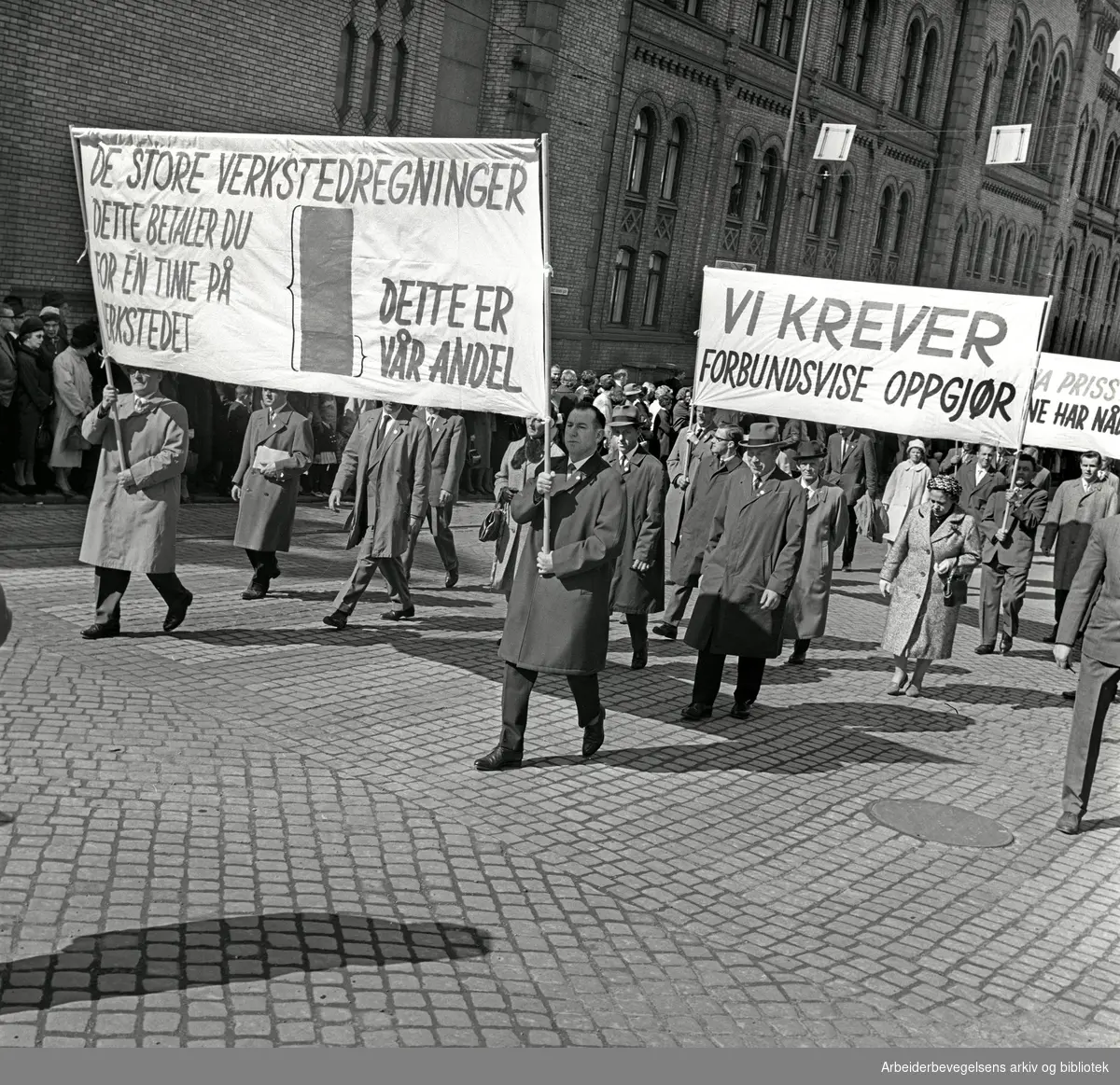 1. mai 1964 i Oslo.Demonstrasjonstoget i Karl Johans gate.Parole: De store verkstedregninger.Dette betaler du for en time på verkstedet (tegning).- dette er vår andel.Parole: Vi krever forbundsvise oppgjør
