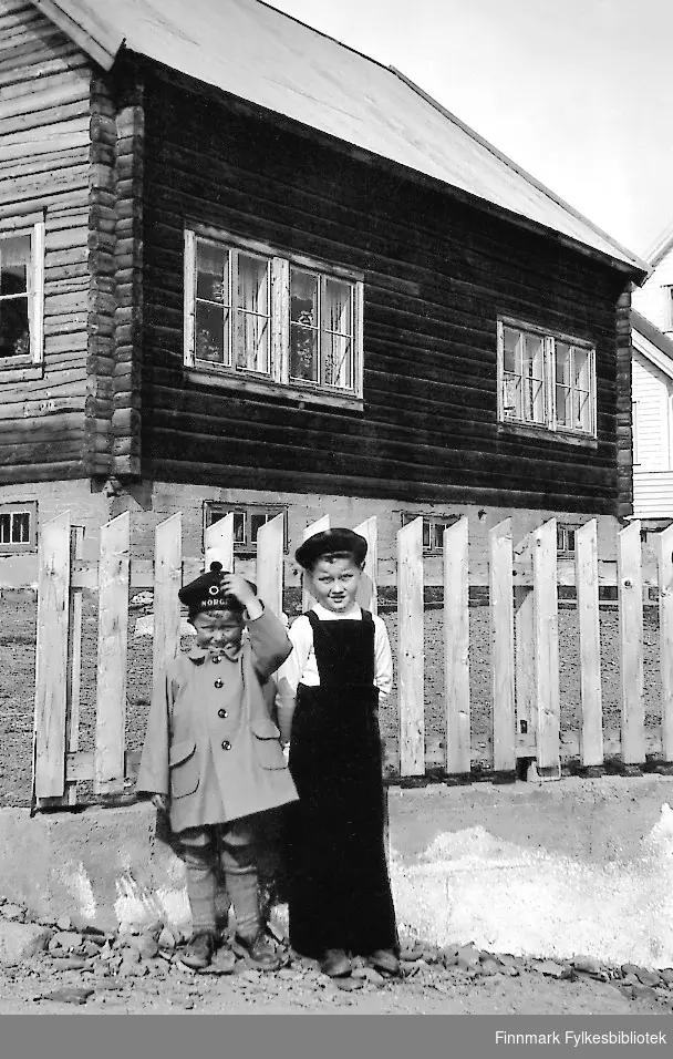 To gutter står på gate ved en tømmerbyggning i Vadsø. Huset tilhører Gjerdrud Betten og gutten til høyre er hennes sønn Bjørn Ottar Betten. Den andre gutten heter Rudolf Betten.