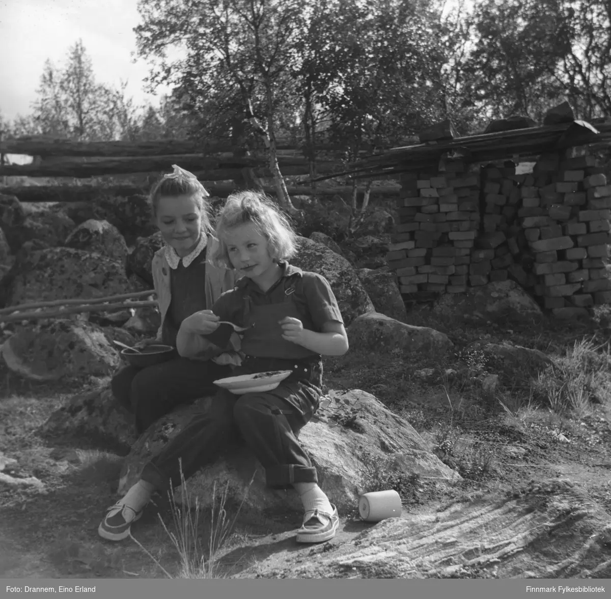 Maija Hoikka (født Gerasimoff)og Turid Lillian Karikoski (Drannem) sitter på en stein og spiser bær i Luolajärvi. I bakgrunn ser man en haug med murstein.