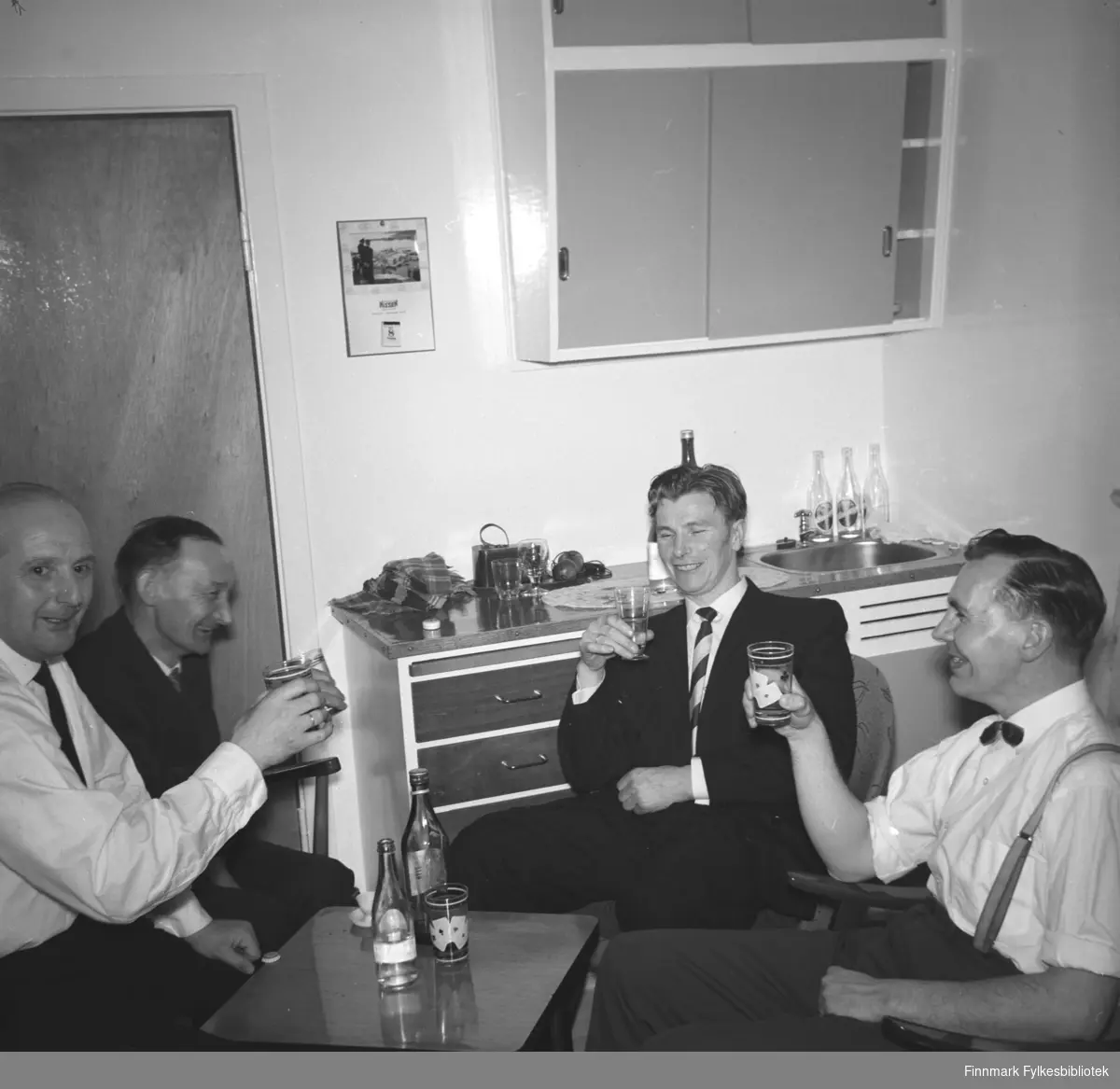 Eino Drannem fotografert i kjøkkenet sammen med sine kompiser. Alle sitter rundt et litet bord og har et glass i hånden og smiler