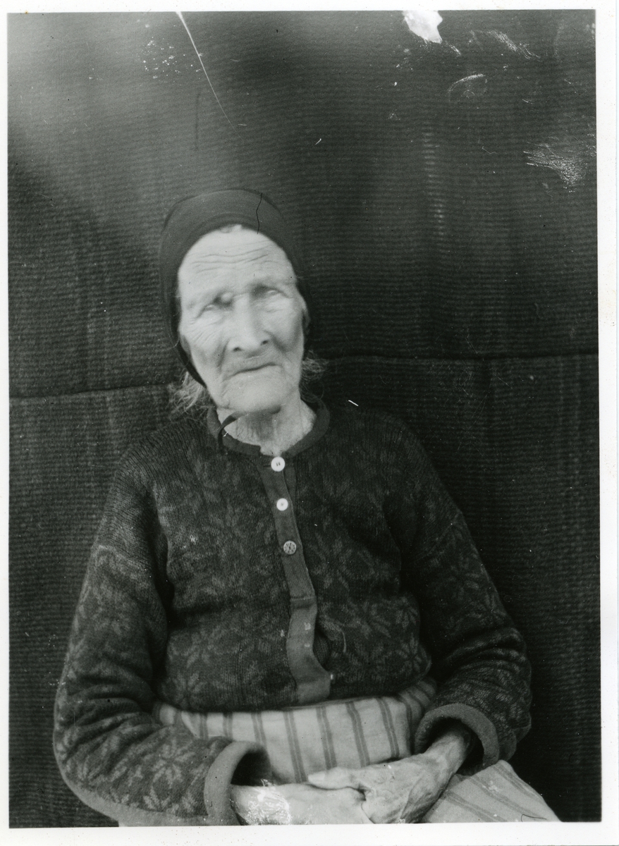 Portrett av en eldre kvinne sittende foran en vegg. Kvinnen er iført et skaut samt en kofte.