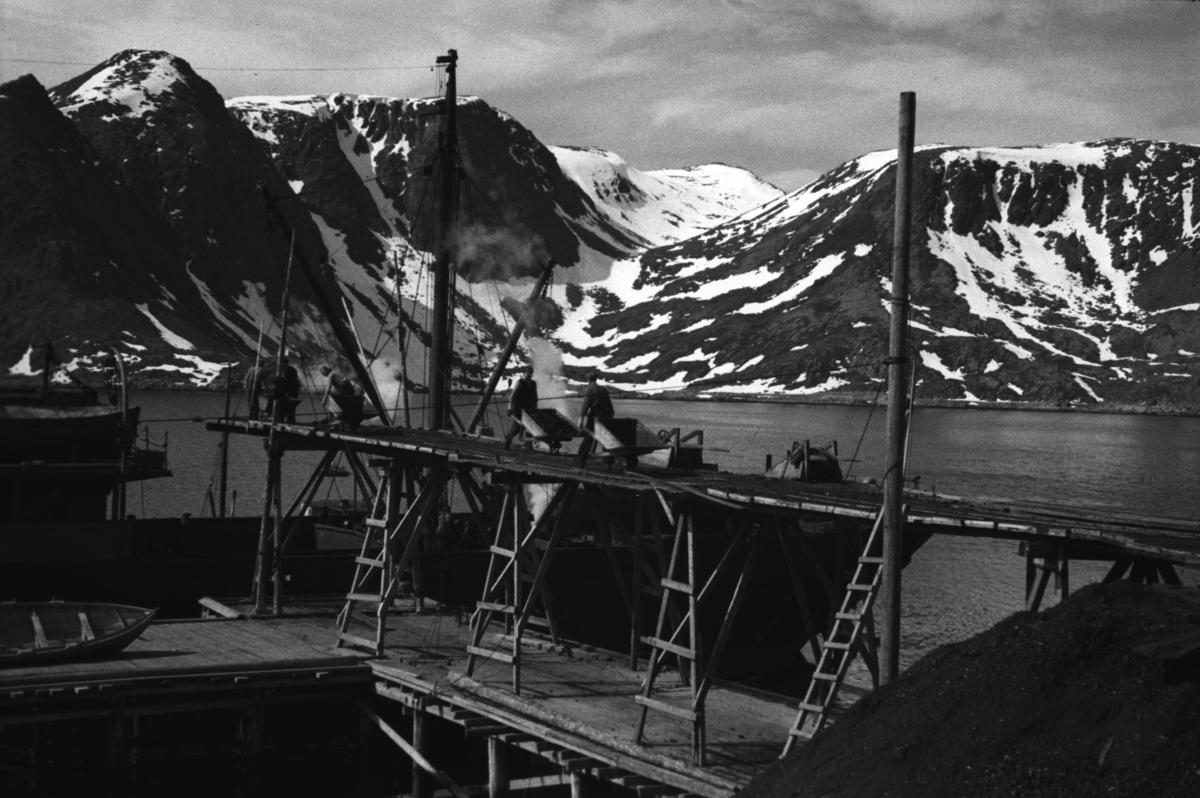 En stor båt ligger ved et uferdig kaianlegg i Honningsvåg, sannsynligvis våren 1946.

Arkitekt Ola Hanche-Olsen arbeidet ved Brente Steders Reguleringskontor i 1946. Hovedadministrasjon for gjenreisning av Nord-Troms og Finnmark ble lagt til Harstad og fikk navnet Finnmark kontoret. Landsdelen Nord-Troms og Finnmark blev oppdelt i syv distrikt med hver sin administrasjon. Honningsvåg, distrikt IV, skulle betjene Nordkapp, Lebesby, Porsanger og Karasjok kommune.

Ola Hanche-Olsen har tatt bildene. Han var født 13. mars 1920 i Borre, død 11. februar 1998 i Gjettum. Han var både arkitekt og barnebokforfatter. Han hadde artium fra 1939, arkitekteksamen fra NTH 1946 og arbeidet deretter ved Finnmarkskontoret 1946–48 før han etablerte egen arkitektpraksis. Han debuterte som barnebokforfatter i 1974 med lettlest-boka "Knut og sjørøverne", og skrev i alt 12 bøker. Han var XU-agent 1944-45, og var også en aktiv fjellklatrer og friluftsmann. Ola var gift med Solveig Hanche-Olsen (f. Falkenberg); de fikk 3 barn, blant dem matematikeren Harald Hanche-Olsen.


