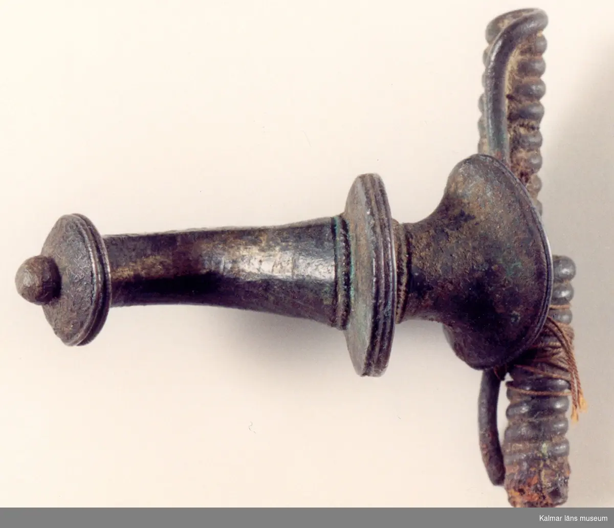 KLM 15. Bågspänne. Armborstfibula av brons. Fibulan har profilerad båge och en liten knopp i ena änden. Nålhållaren något avbruten. Datering: äldre romersk järnålder.