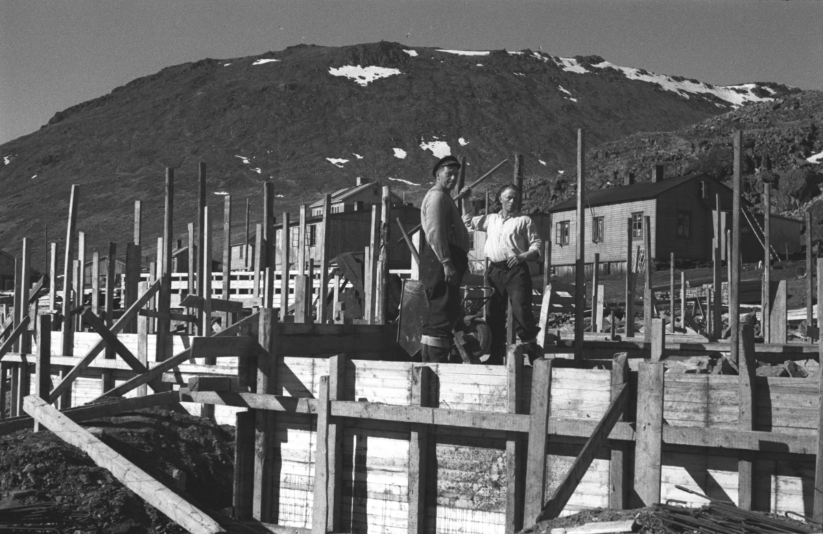 Forskaling til en grunnmur er satt opp i Honningsvåg etter andre verdenskrig og to menn med en trillebår fotografert inne i byggverket.

Arkitekt Ola Hanche-Olsen arbeidet ved Brente Steders Reguleringskontor i 1946. Hovedadministrasjon for gjenreisning av Nord-Troms og Finnmark ble lagt til Harstad og fikk navnet Finnmark kontoret. Landsdelen Nord-Troms og Finnmark blev oppdelt i syv distrikt med hver sin administrasjon. Honningsvåg, distrikt IV, skulle betjene Nordkapp, Lebesby, Porsanger og Karasjok kommune.

Ola Hanche-Olsen har tatt bildene. Han var født 13. mars 1920 i Borre, død 11. februar 1998 i Gjettum. Han hadde artium fra 1939, arkitekteksamen fra NTH 1946 og arbeidet deretter ved Finnmarkskontoret 1946–48 før han etablerte egen arkitektpraksis. Han debuterte som barnebokforfatter i 1974 med lettlest-boka "Knut og sjørøverne", og skrev i alt 12 bøker. 

Han var XU-agent 1944-45, og var også en aktiv fjellklatrer og friluftsmann. XU var den største og viktigste allierte etterretningsorganisasjonen i det okkuperte Norge under andre verdenskrig. Det meste av XUs virksomhet ble holdt hemmelig til 1988. Ola var gift med Solveig Hanche-Olsen (f. Falkenberg); de fikk 3 barn, blant dem matematikeren Harald Hanche-Olsen. 


