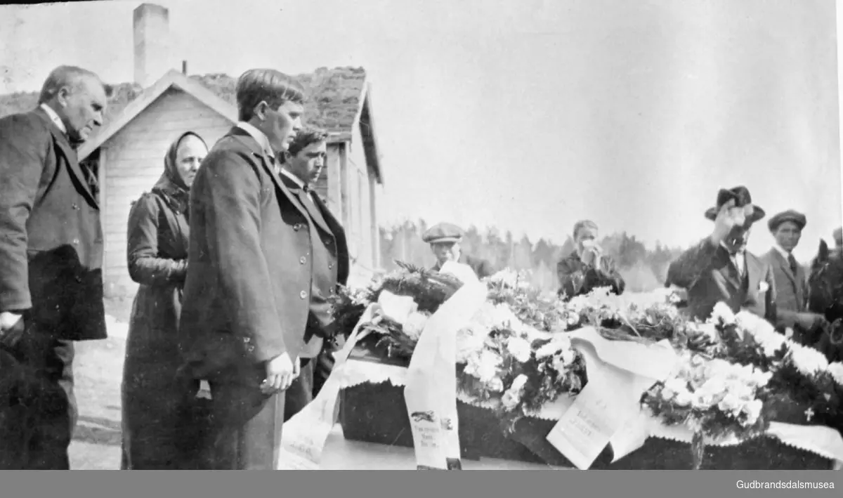 Marit Nordslettens begravelse. Åtte personer ved kiste, kiste med blomsterkranser oppå, en person holder en hest, bygning i bakgrunnen.