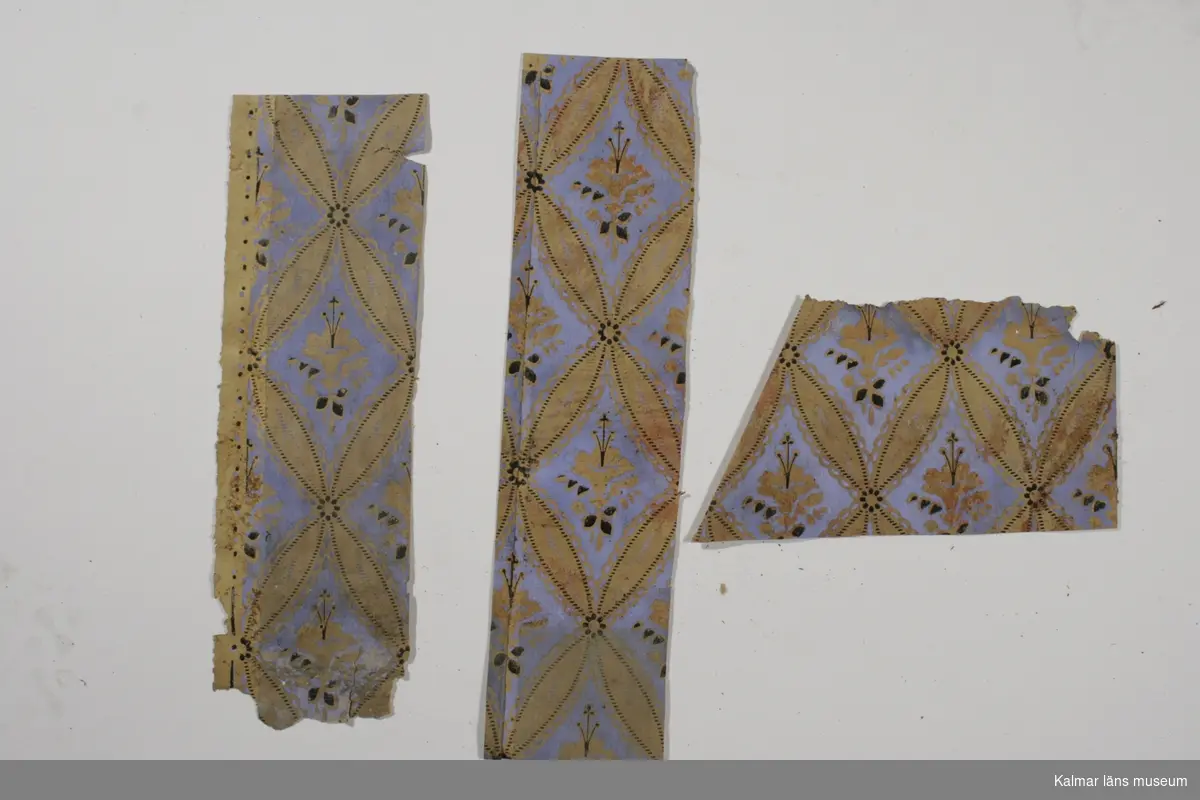 KLM 39224:3. Tapet av papper. 3 bitar. Tapeten har blålila botten med diagonalrandigt mönster i guld som ramar in stiliserade blommor i guld och svart. Datering: ca 1860-1870-tal.