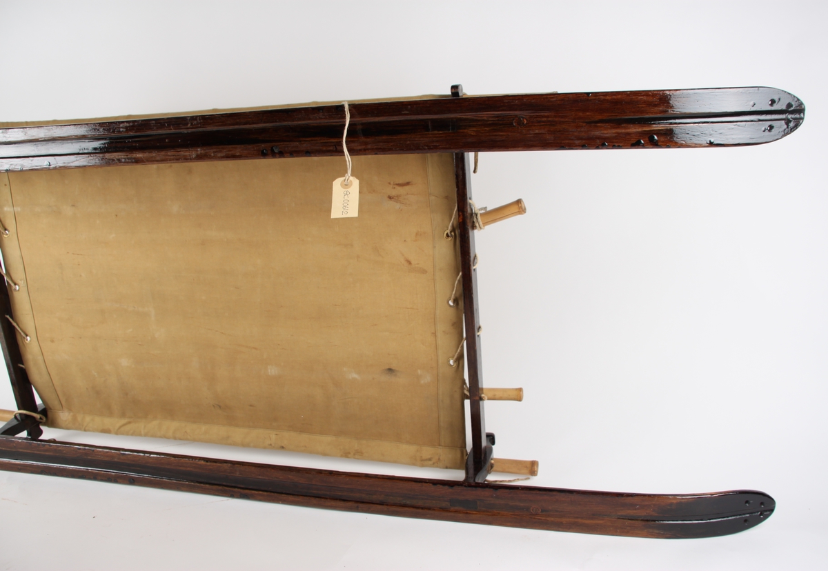 Skikjelke for en person med meier i tjærebeiset tre. Pulkens "sete" er av seilduk, spent over en ramme av blant annet bambusstenger.