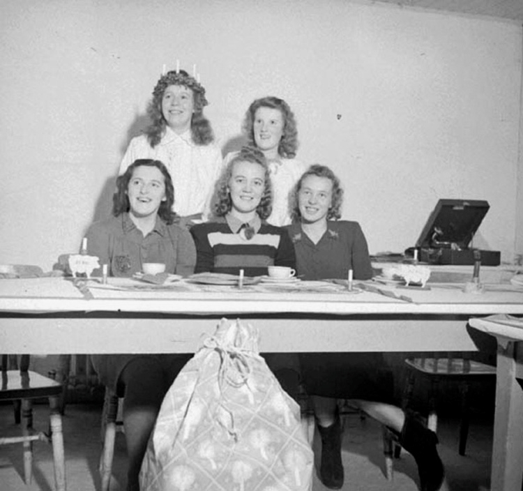 Fem flickor vid ett bord med en vevgrammofon i bakgrunden. Den ena flickan är klädd i vitt Lucialinne och har ljuskrona på huvudet. På bordet står kaffekoppar och små ljus och framför bordet en julklappssäck.