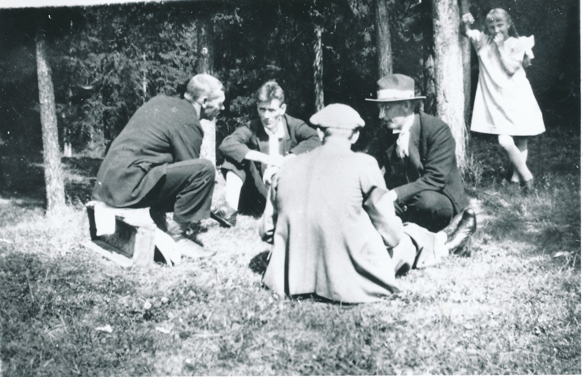 Fire menn sitter og spiller kort i skogen iført søndagsantrekk. En av de sitter på en kasse. Jente i 10-12-årsalderen ikledd kjole og med flettet hår står og ser på.