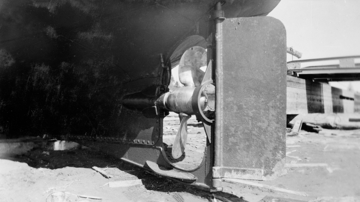 Detalj fra akterstavnen på Glomma fellesfløtingsforenings fartøy «M/S Askeladden». Fotografiet er tatt på skrå bakfra, mot propellen og roret, på et tidspunkt da fartøyet var trukket på land, antakelig for skifte av propell. Bildet ble antakelig tatt i 1941. Askeladden ble bygd ved Glommen mek. Verksted i Fredrikstad i 1925 etter de samme tegningene som den tre år eldre slepebåten «Per». Skroget på Askeladden var 34 fot langt og 10,8 fot bredt. Fartøyet ble brukt i bukseringsarbeid i den nedre delen av Glomma. Askeladden gikk i Glomma fellesfløtingsforenings tjeneste så lenge det pågikk fløting i dette vassdraget.
