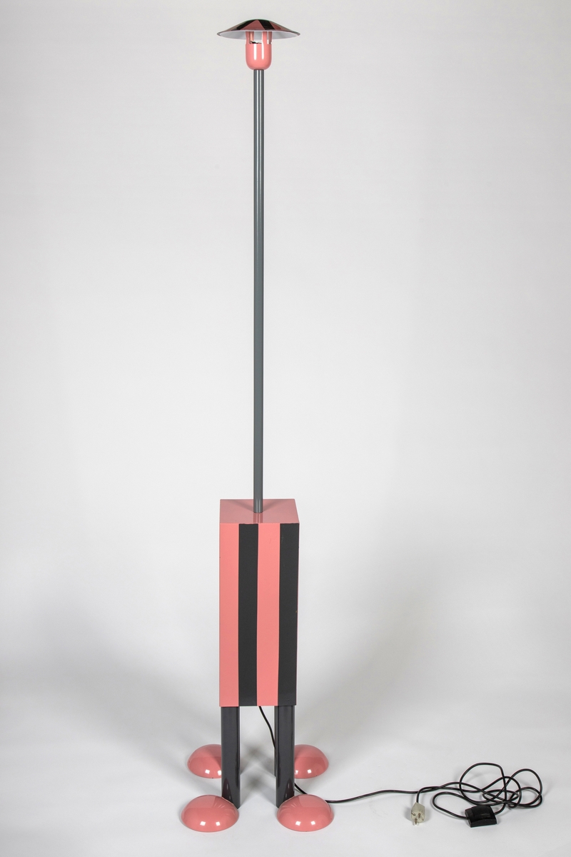 Skulpturell stålampe med antropomorfisk uttrykk - øverst lyspæresokkel med liten skjerm over til spredning av lys (hode med hatt), festet til langt stålrør (hals) på kasse (kropp) over fire bein med rosa "sko" vendt til hver sin side. Kasse og skjermhatt har vertikale striper i grått og rosa.