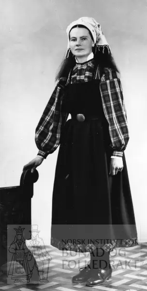 Kvardagskledd kvinne frå Øvre Hallingdal. Ho har ruta skjorte med kvit skjorte under, livjul og kvitt tørkle med mønster på hovudet.