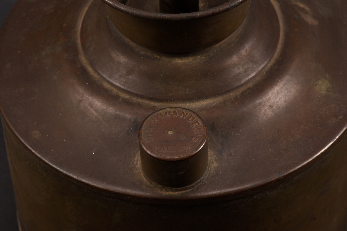 Cylindrisk gasoljelampa tillverkad av mässing. I mitten en cylindrisk vekhållare som är fäst i gängat påfyllnadslock. På sidorna av oljehuset sitter två cylindriska upphöjningar med oklar funktion. Den ena är öppningsbar och locket har ursprungligen varit förankrad i oljehuset via en länk.