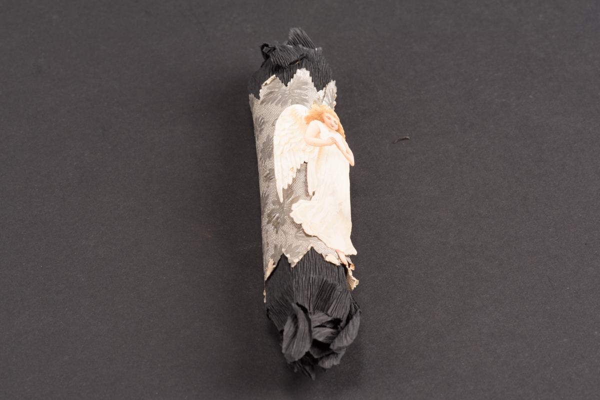 Rund begravningskaramell inlagd i svart kräppapper. Ett silverfärgat papper med uddig kant och inpräglade blommor sitter runt karamellen. Ovanpå detta sitter ett bokmärke i form av en ängel.