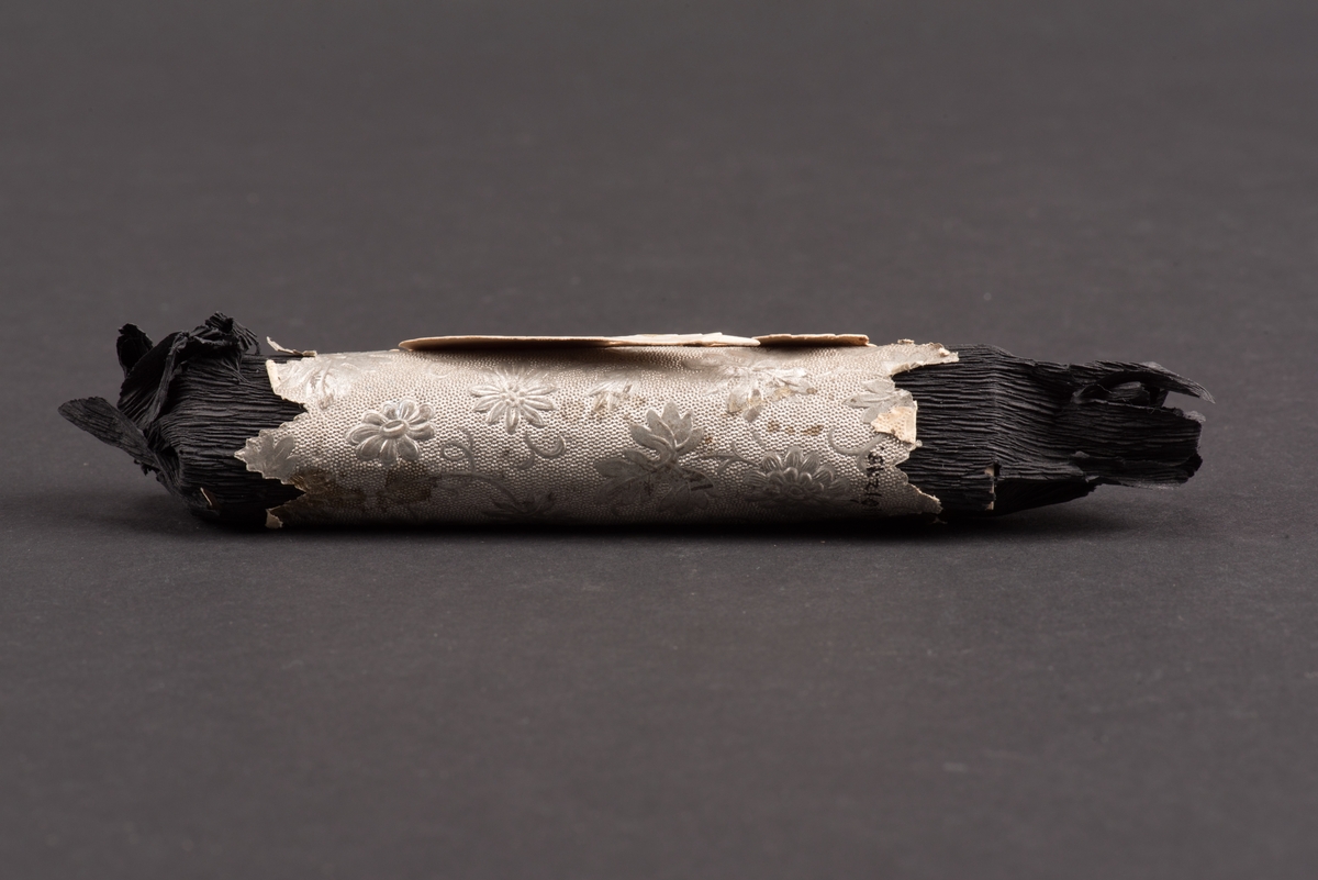 Rund begravningskaramell inlagd i svart kräppapper. Ett silverfärgat papper med uddig kant och inpräglade blommor sitter runt karamellen. Ovanpå detta sitter ett bokmärke i form av en ängel.