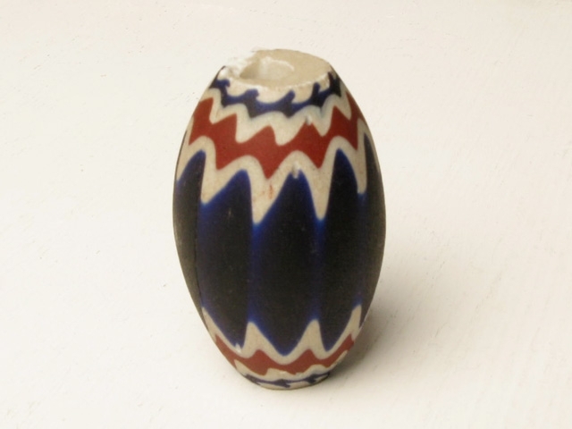 Halssmycke; berlock av målad porslin. I färgerna: blått, vitt och rött. Äggformad och med ett stort och flera små hål rakt igenom.