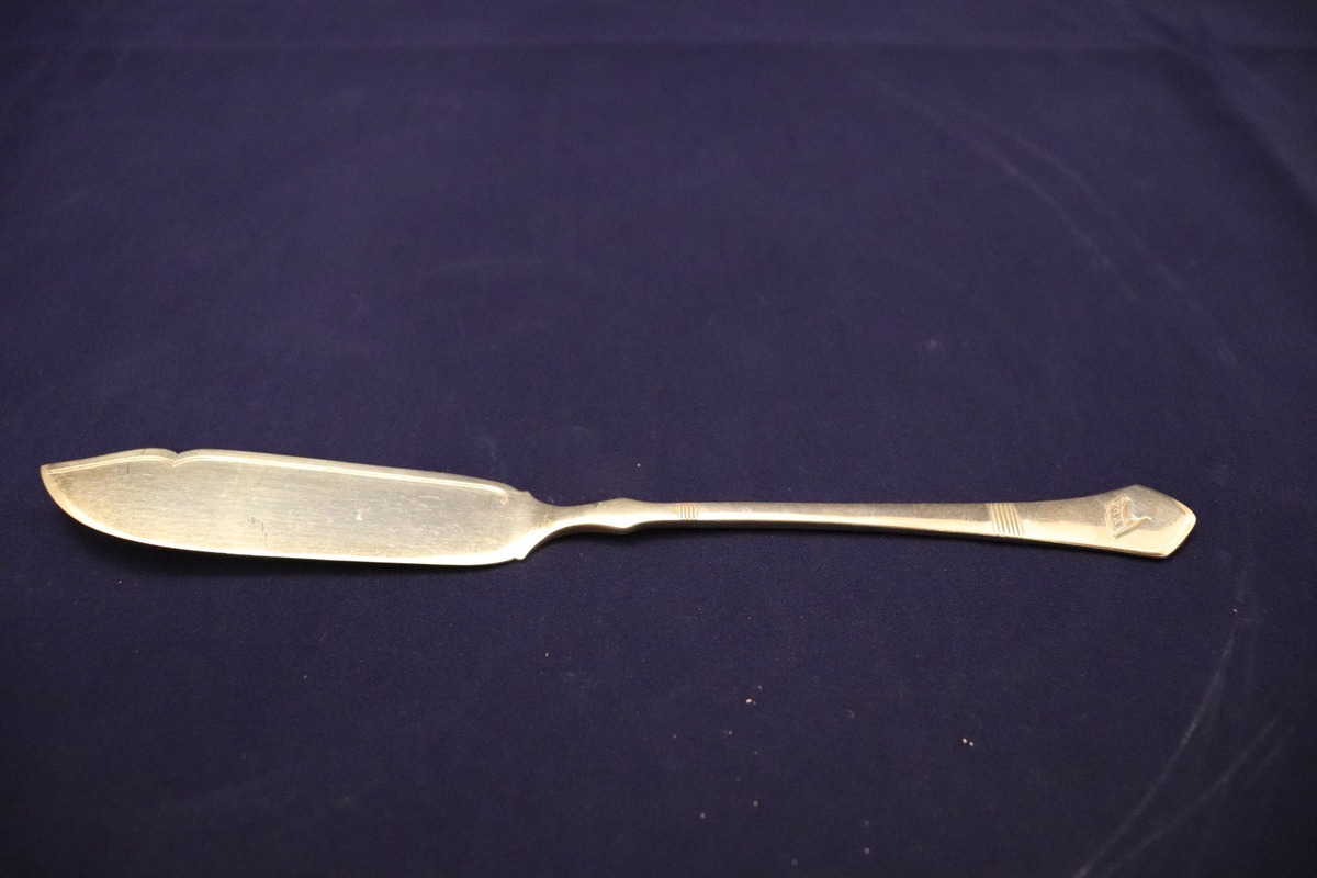 Kniv brukt til servering av fisk, trolig ombord i VDS sine hurtigruteskip. Kniven har den typiske spatel-formen for lettere å skille filet fra fiskebeinet. Rederiflagget til Vesteraalens dampskibsselskap er stemplet inn i skaftet av kniven, i tillegg er det skåret inn enkle dekorasjonsdetaljer i skaftet. På underside av skaftet er det stempler som viser til at kniven er sølvplett, samt et produksjonsstempel.
