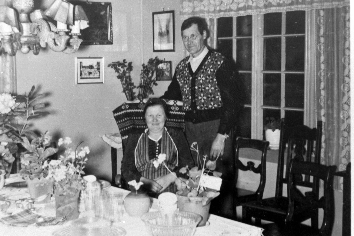 Interiør fra stue. Kvinne sitter ved bord med blomster og presanger, ektemannen står bak.