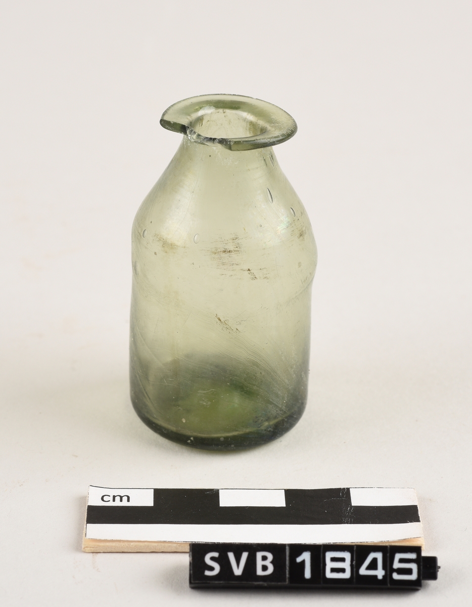 Sylindriskformet glassflaske. Deler av randen mangler. På toppen av flasken er det et sirkelformet hull. Hullet har en diameter på 1,2 cm. Flasken er lys grønn i farge.