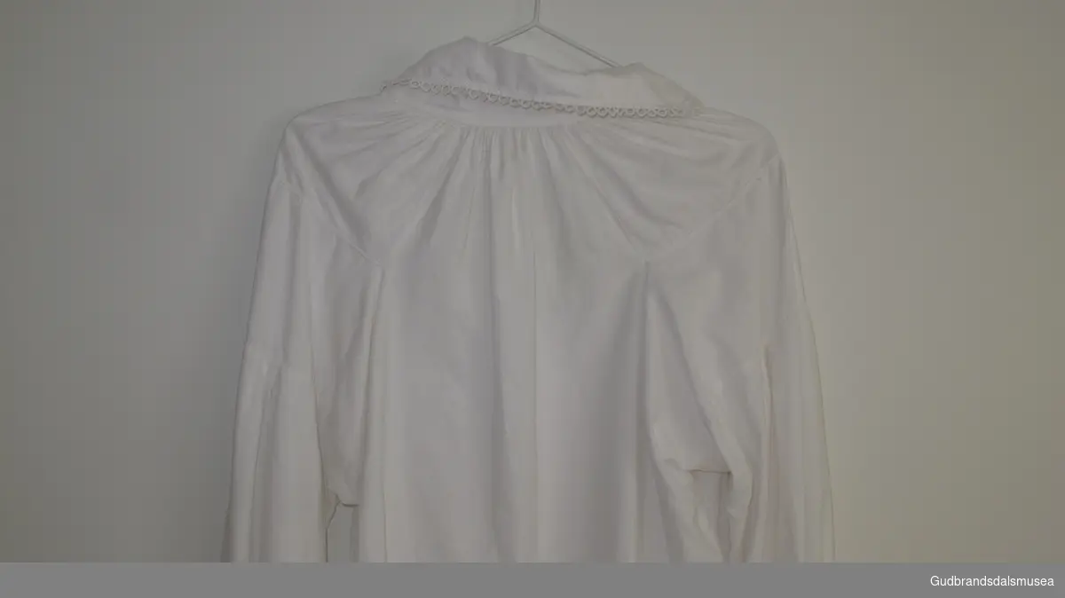 Bunadsskjorte sydd i hvitt bomullsstoff. Kniplinger (nuppereller) og kjedesøm på krage og ermer. Delvis håndsøm, delvis maskinsøm. Fillete på kragen.