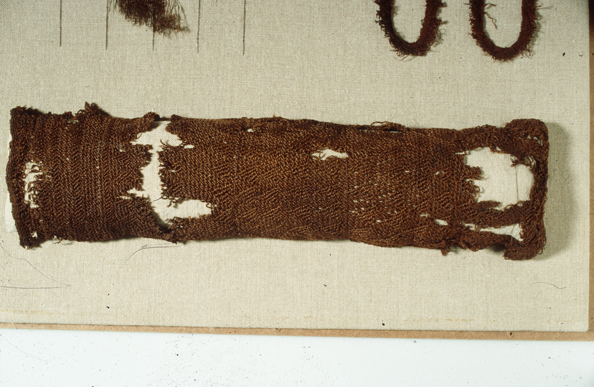 Stykke av sprangvevet hoselegg av ull med vevkant, ca. 1,5 x 11,5-cm. 